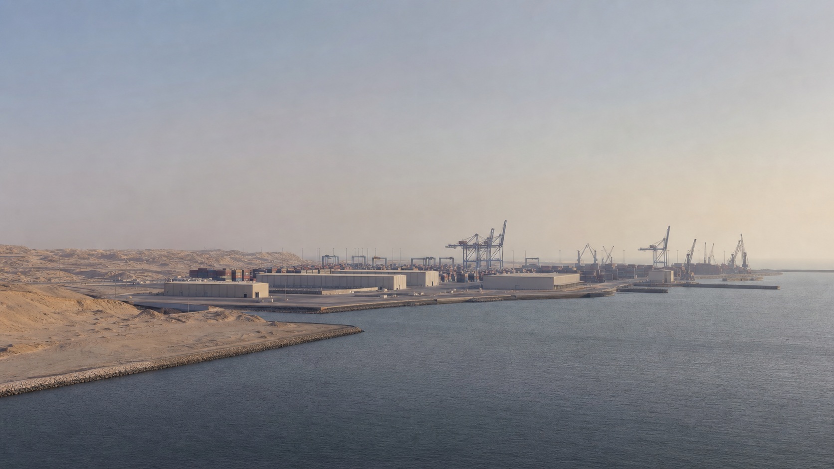 Calm arid coastal port with low warehouses, distant cranes, container stacks, and open water.